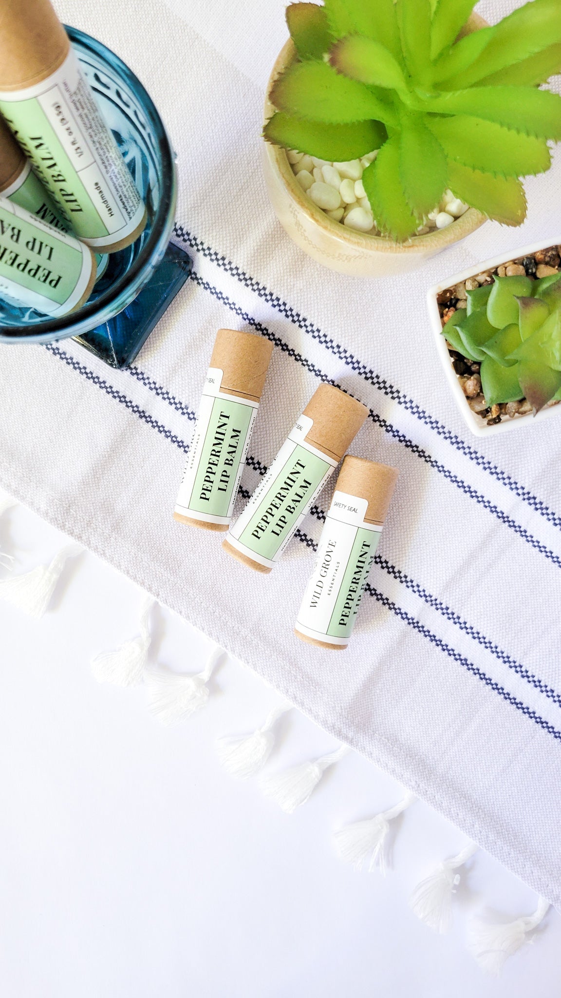 Peppermint lip balms in cardboard tubes with a green label being displayed in a glass cup and on a hand towel with plants in the background.