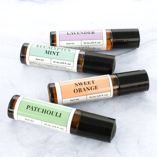 4 essential oil roll-on bottles each in 10ml amber bottle on a marble backdrop in Patchouli, Sweet Orange, Eucalyptus Mint, and Lavender scents.
