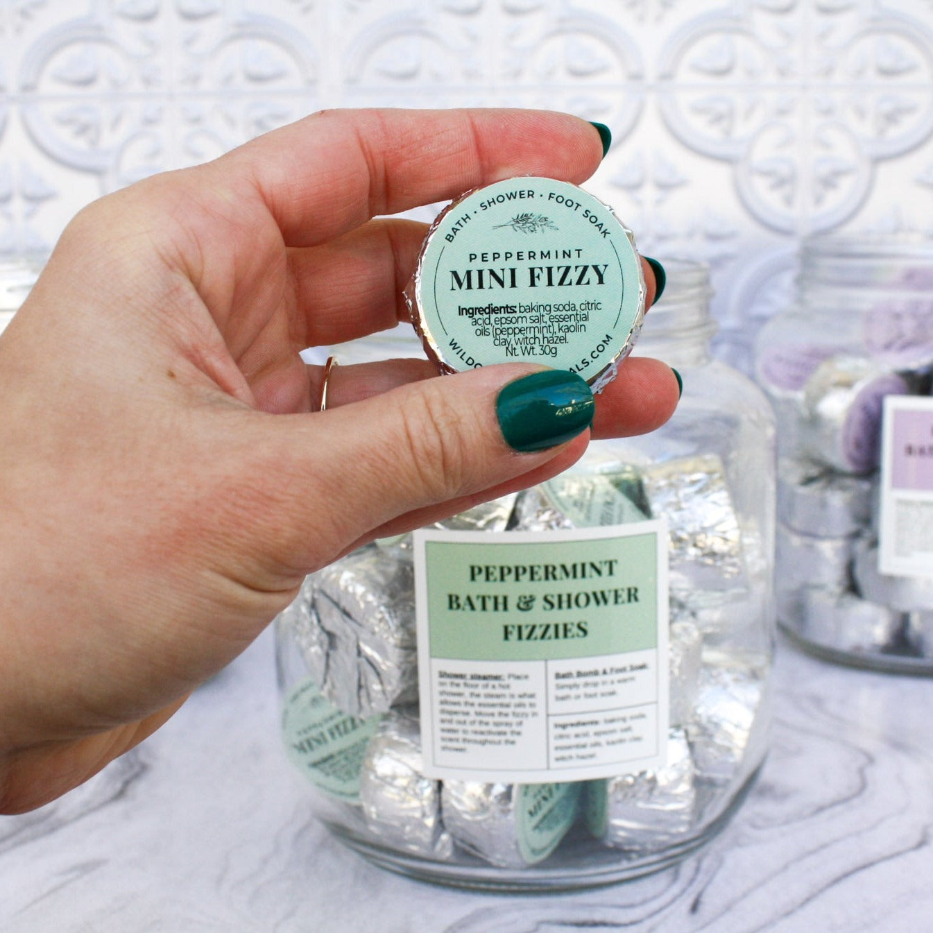 Peppermint scented mini bath and shower fizzy held in front of a jar full of fizzies wrapped in foil with green labels.