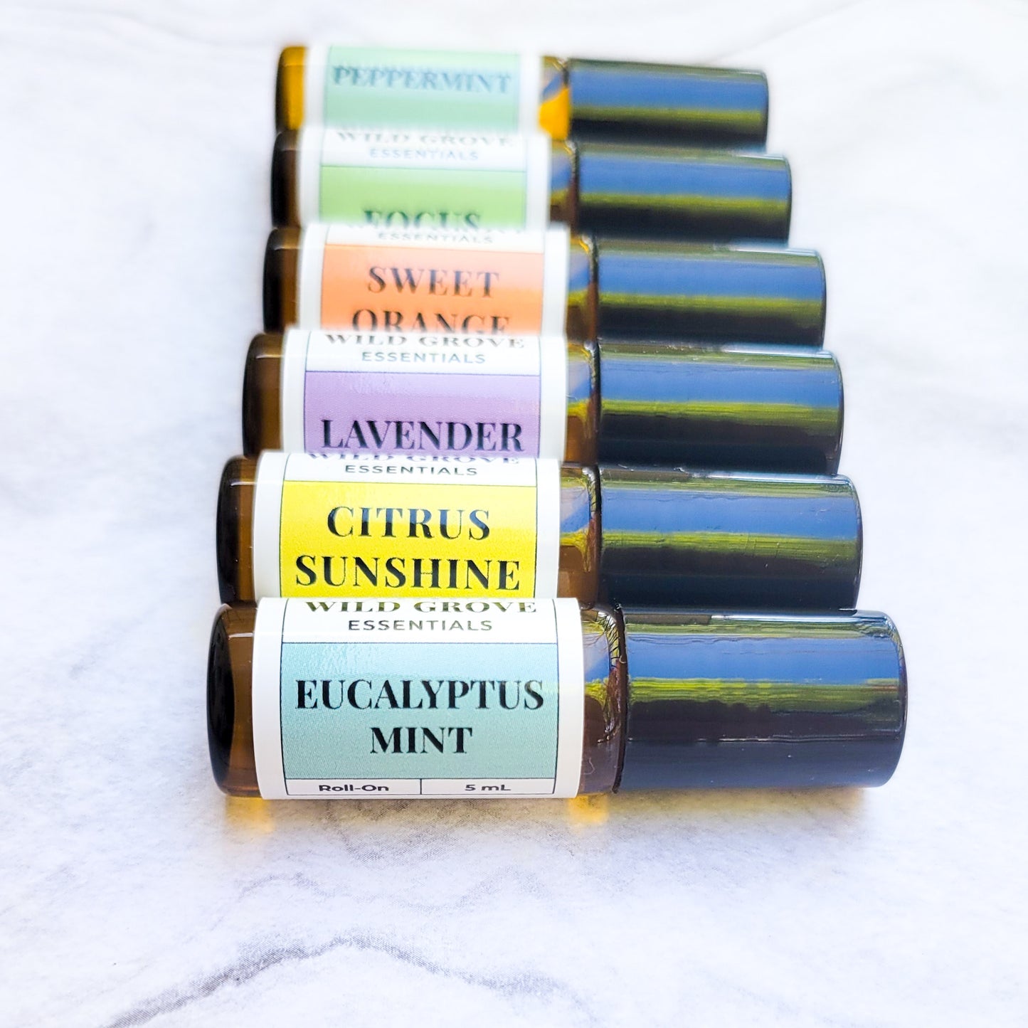 Mini essential oil roll ons in small 5ml bottles lined up in Eucalyptus Mint, Citrus Sunshine, Lavender, Sweet Orange, Focus, and Peppermint scents laid on a marble backdrop.
