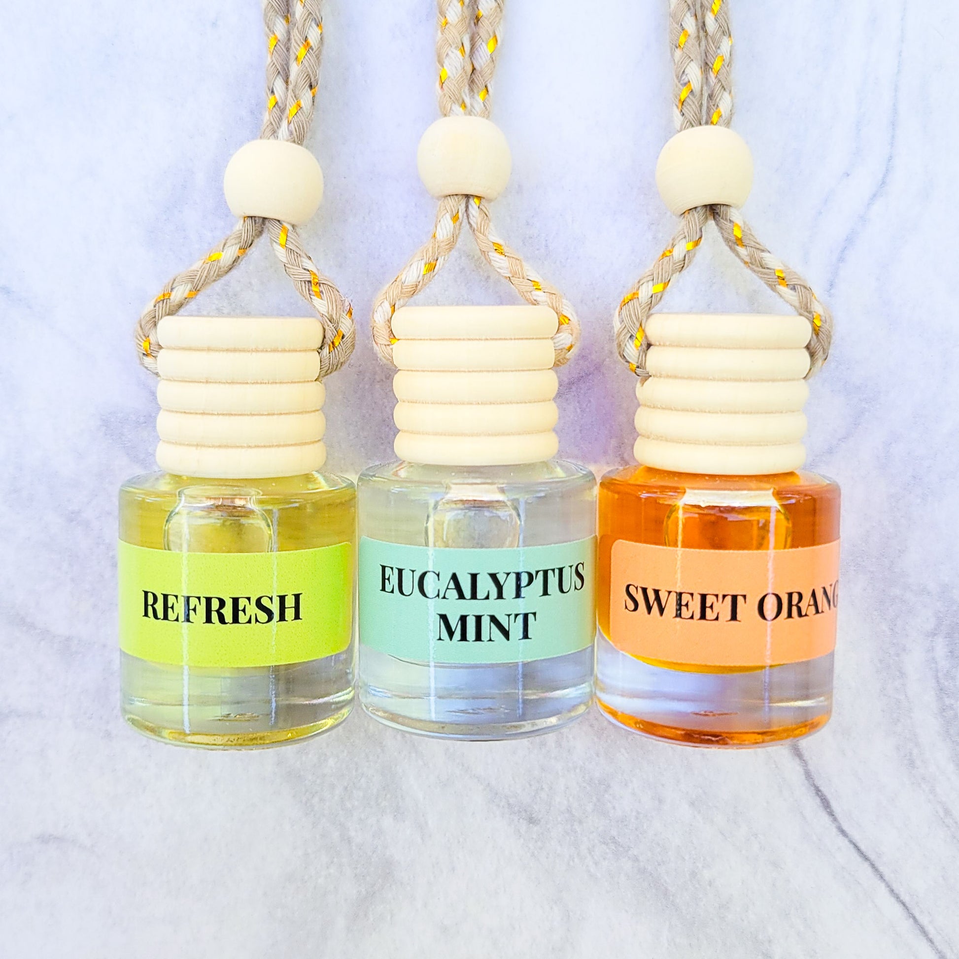 Car fresheners in glass bottles with wooden tops  in Eucalyptus Mint, Sweet Orange, and Refresh scents laid on a marble backdrop.